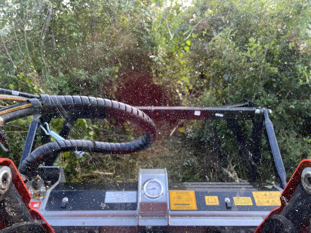 View from the operator's seat through a debris-covered windscreen, the mulching head cutting into green brush